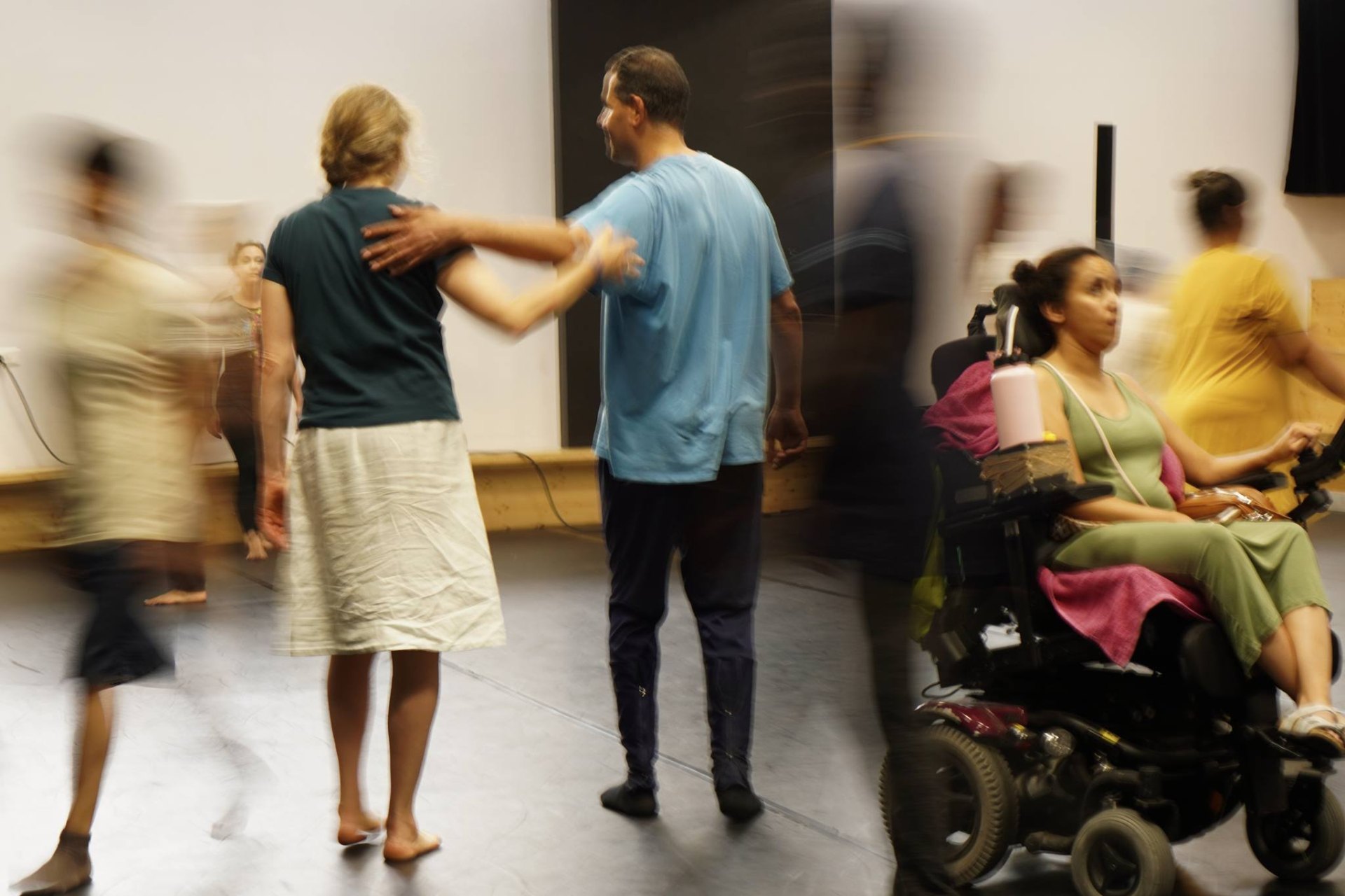 Das Foto ist in Bewegung aufgenommen, deshalb sind die Menschen verschwommen. Fünf Menschen laufen durch das Foto, eine Person fährt mit dem Rollstuhl am rechten Rand aus dem Rahmen. Die beiden in der Mitte haben die Arme umeinander gelegt.