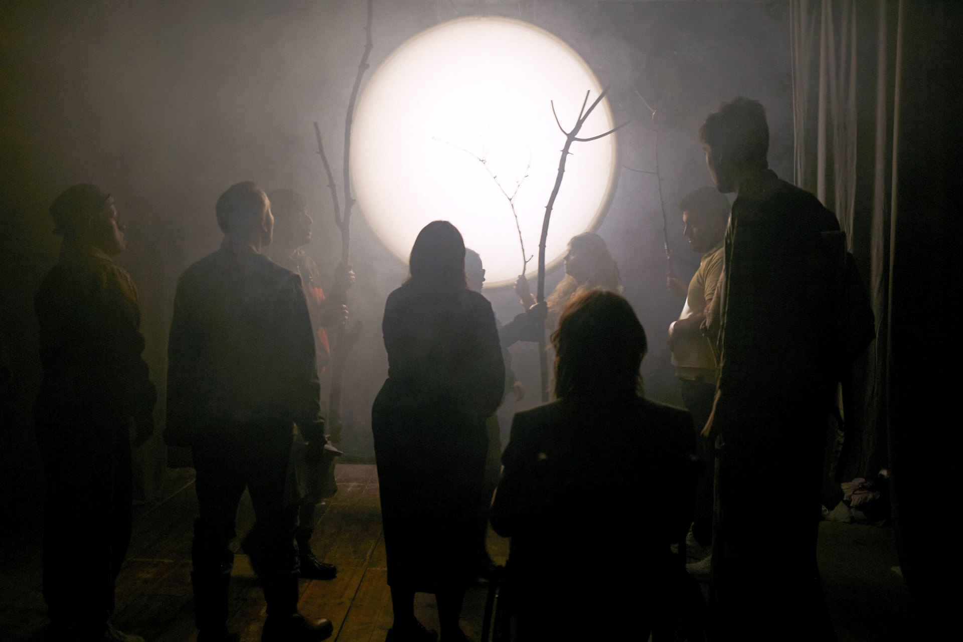 Neun Leute in nebligem Raum, an der Rückwand ist ein Vollmond projiziert. Alle sind dem Mond zugewandt, mehrere halten lange, kahle Stöcke in der Hand.