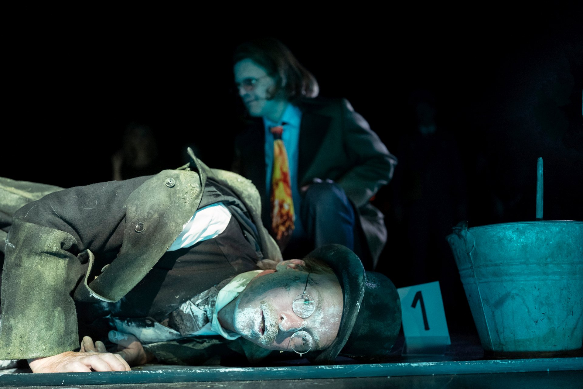 Im Vordergrund liegt ein Performer mit Zylinder und kreisrunder Brille auf dem Boden. Neben ihm steht ein Schild mit "1", wie bei Beweisstücken. Rechts steht ein Metalleimer. Im Hintergrund kniet ein Mensch mir rot-gelber Krawatte.