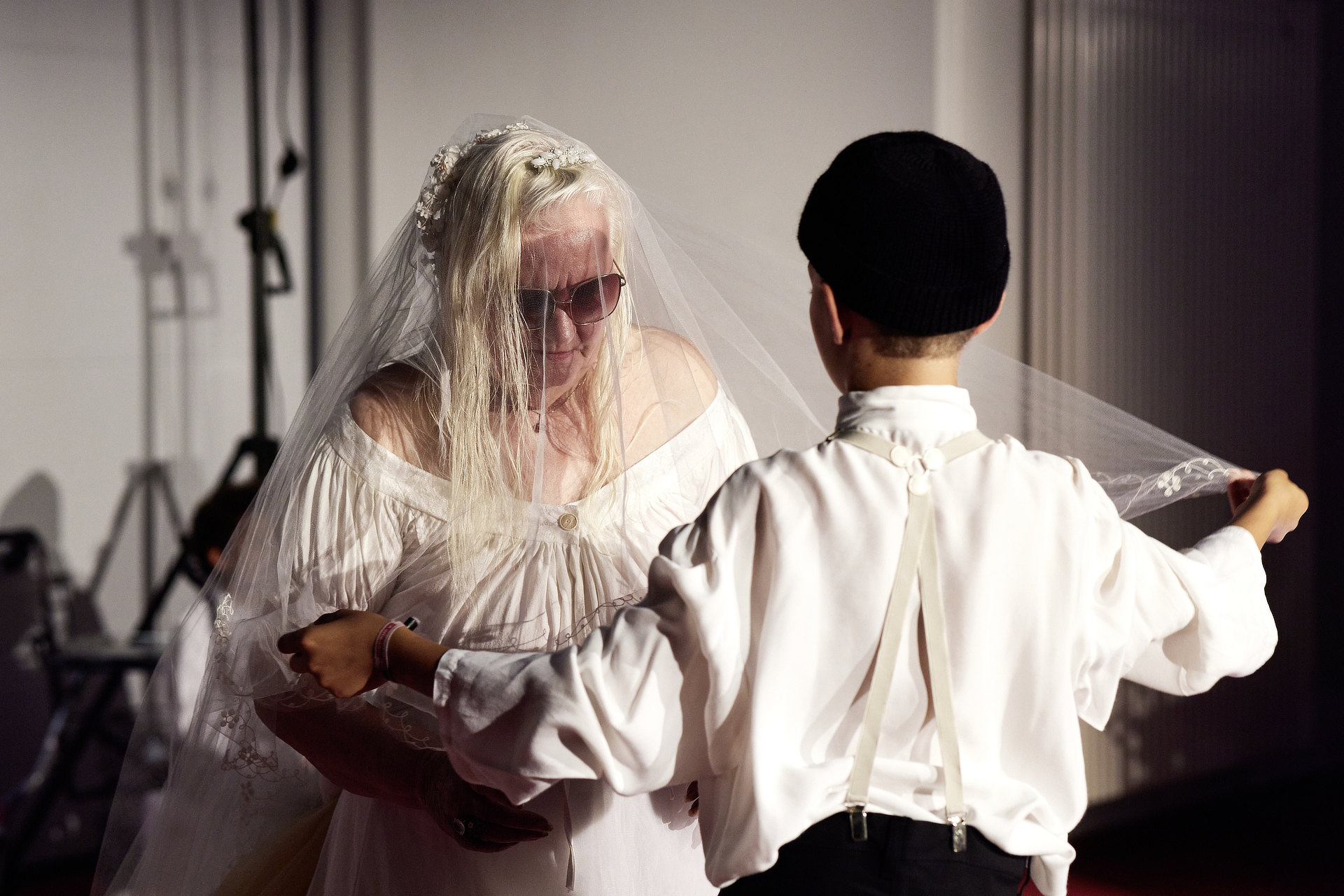 Zwei Performer stehen sich gegenüber. Die vordere Person steht mit dem Rücken zur Kamera. Sie trägt weißes Hemd und eine schwarze Mütze. Die hintere Person trägt ein weißes Kleid, einen Schleier und Sonnenbrille.