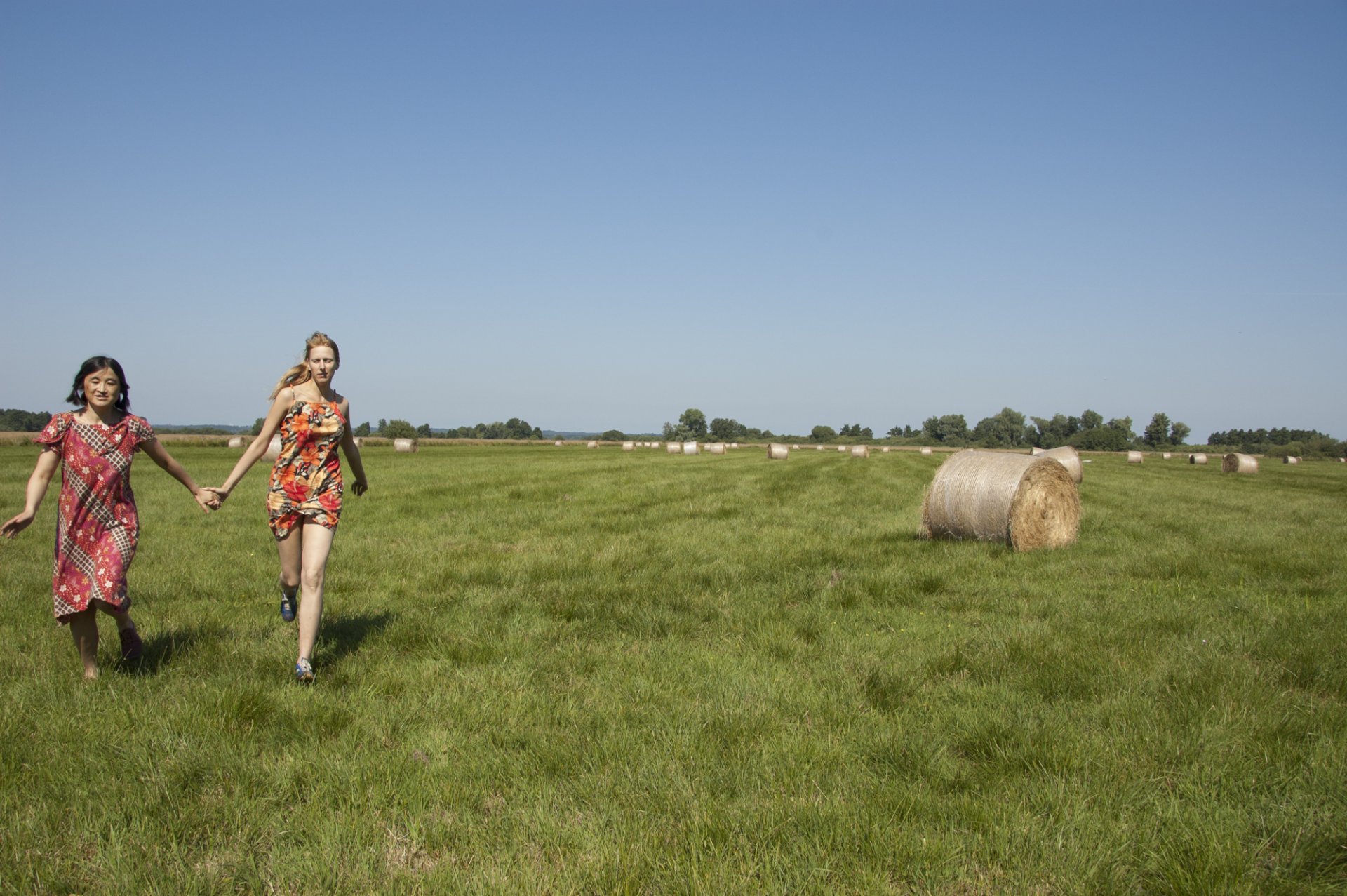 Das Foto zeigt eine satt-grüne Wiese, blauen Himmel und rechts einen Heuballen. Links springen zwei Personen über die Wiese, sie halten sich an den Händen.
