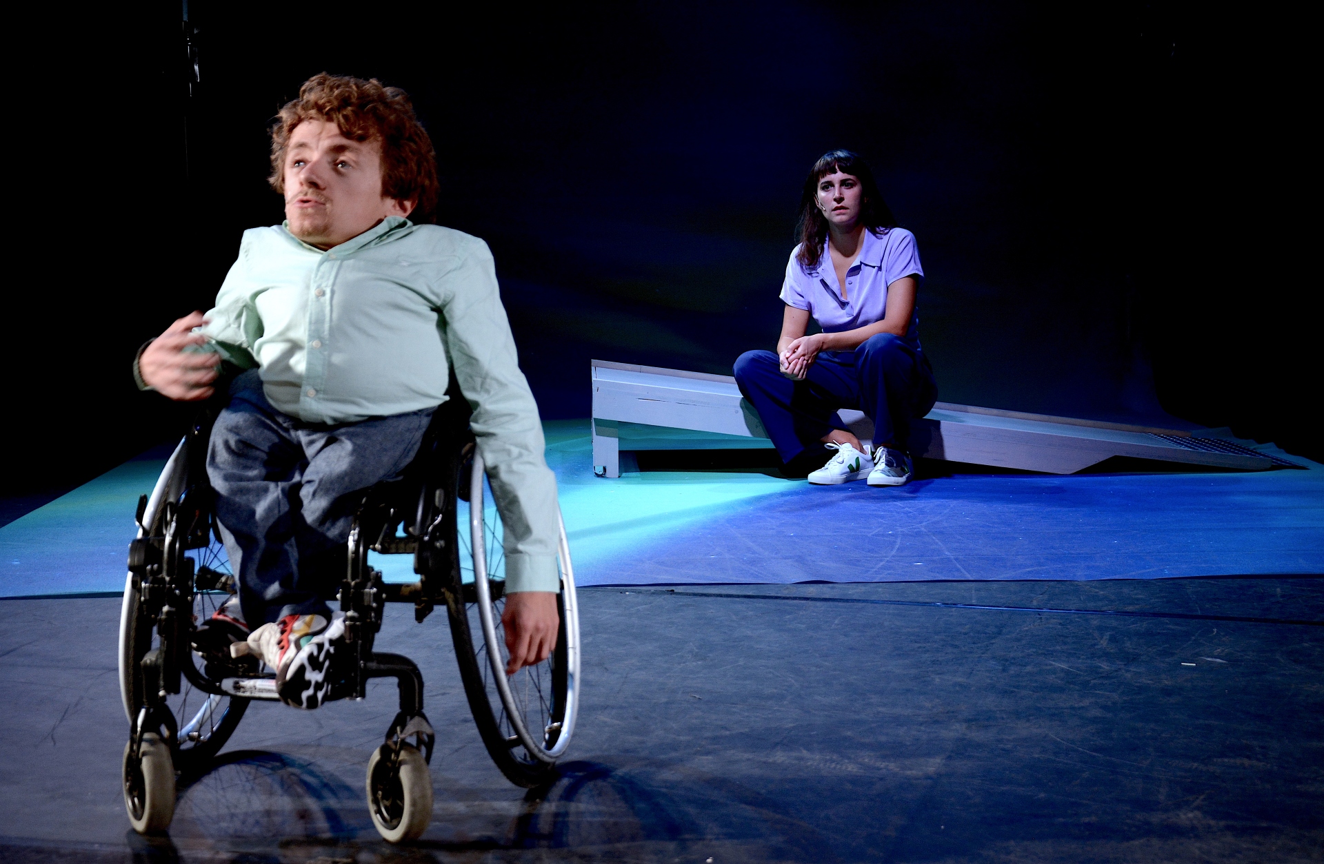 Im Bildvordergrund links spricht eine Person, die ein Hemd trägt und einen Rollstuhl nutzt, zum Publikum. Im Bildhintergrund rechts sitzt eine Person auf einer Rampe und sieht den Menschen vor ihr aus der Distanz an.