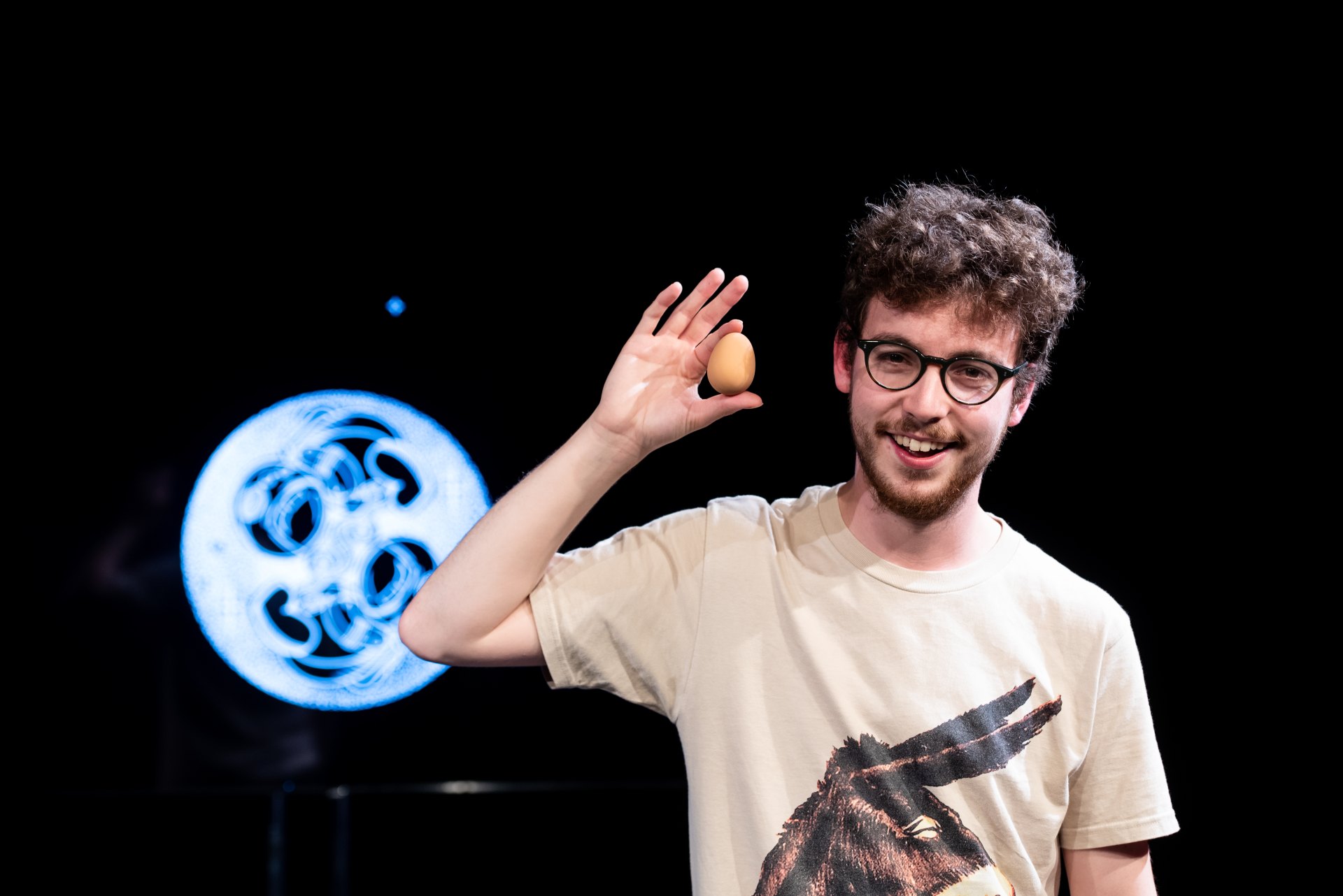 Das Foto zeigt eine Person mit Brille, die ein Ei in der rechten Hand neben ihren Kopf hält. Der Hintergrund ist schwarz, bis auf eine kreisrunde LED Lampe, in deren Mitte unregelmäßige Wirbel leuchten.