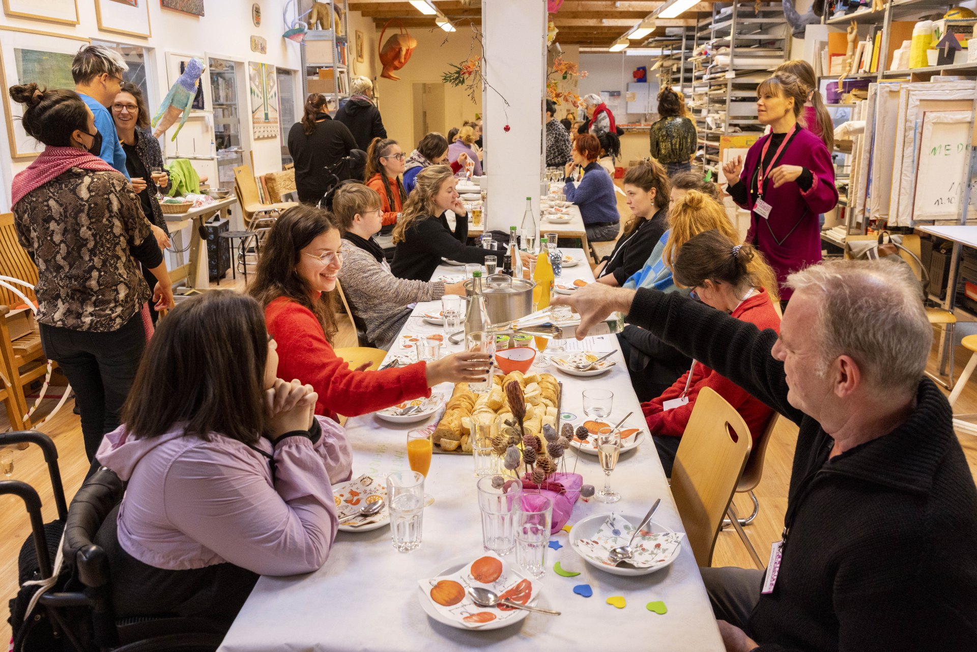 Foto von einer reich gedeckten Tafel, an der viele Menschen sitzen. Auf dem Tisch befinden sich Getränke und Snacks. Im Raum sind viele Kunstmaterialien zu sehen, zum Beispiel Leinwände.